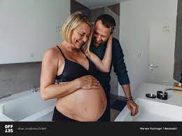 Happy mature pregnant couple watching expectant mothers belly in bathroom  stock photo - OFFSET