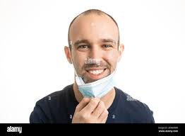 A Caucasian male with a blue t-shirt and stubble facial hair pulling down  his blue surgical face mask to smile. White background Stock Photo
