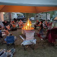 A group of people gathered around a campfire on a lawn, under a large canopy, with a dog, chairs, and some homes in the background.
