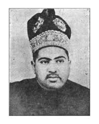 Photograph of Sir Sadiq Muhammad Khan Abbasi V, Nawab of Bahawalpur,  standing next to a chair, facing the camera. He wears a richly embroidered  robe, slipper and a decorative headdress. . . . . . . . . . . . . . . . . .  . .