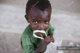 Goree, Senegal- 6 de diciembre de 2017: Retrato de un niño negro mordiendo  una cinta de plástico en la escena callejera y mirando a la cámara — grave,  San Luis