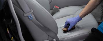 A gloved hand uses a brush to clean a perforated leather car seat where milk was spilled