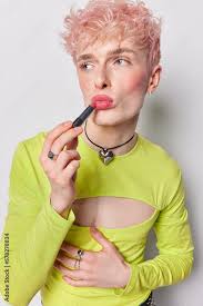 Serious gay applies lipstick has feminine manners non traditionalsexual  orientation wears green fashionable jumper has pink dyed curly hair  prepares for date isolated over white wall. Lgbt community Stock Photo