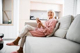 A woman looking forward with her arm on the couch arm and smiling. Smiling Elderly Woman Sitting On Couch And Holding Smartphone Stock Photo Image Of Retired Happy 174156008