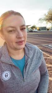VD: Joy is a white woman with brown hair pulled back into a braid. She is  sitting in a parking lot in a wheelchair with the sun coming up behind her.  She is wearing a gray three-quarter zip pull over ...
