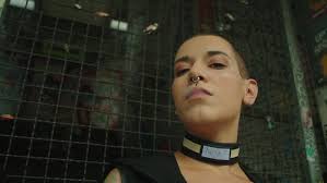 Close up of a woman wearing a nose ring and choker necklace while smoking a  cigarette standing in front of a cage