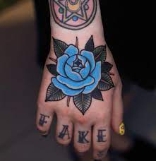 Traditional Style Blue Rose Tattoo On The Right Hand And A Word Fake On The Fingers Rose Hand Tattoo Traditional Hand Tattoo Blue Rose Tattoos