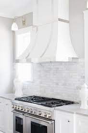 Small white cottage kitchen features a farm sink and a white vent hood with a white vintage stove keeping a light and airy appeal. Tile Backsplash Stops At Kitchen Hood Design Ideas