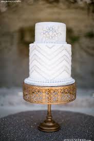 Decorated with layers of sugar tassels, edible gold sugar beads, gold geometric patterns, a gathered fabric detail with edible brooch and topped off with black ostrich feathers and gold beaded decorations. Great Gatsby Inspired Wedding Cake White Wedding Cake With Silver Sparkle Jamilah Photography Pao Art Deco Wedding Cake Silver Wedding Cake Art Deco Cake