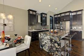 A stepped island creates a dining space showcasing the veining of brittanicca gold. Natural Stone Granite Countertop With Waterfall Down One Side Of Island Olympia Ii Show Home Countertops Granite Countertops Kitchen Ideas New House