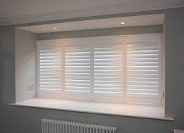 Restful bedroom boasts a glass column lamp placed on a white art deco nightstand in front of a window covered in plantation shutters. White Shutter Blinds White Shutters Essex White Window Shutters