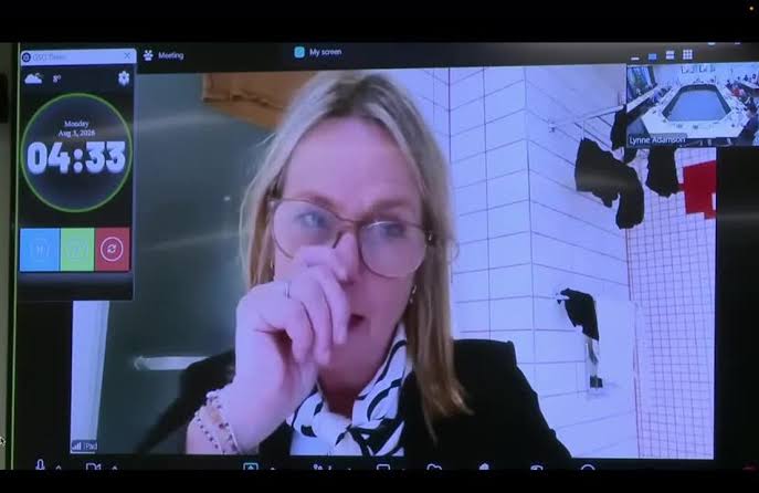 A screenshot of a Zoom council meeting showing a politician participating from a bathroom setting with clothing visible in the background.