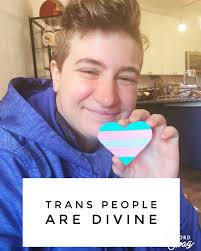 ID: A selfie of Ace holding a heart-shaped patch in the transgender flag  colors. They're grinning and wearing a bright blue sweatshirt. A white box  is underneath the hand holding the heart,