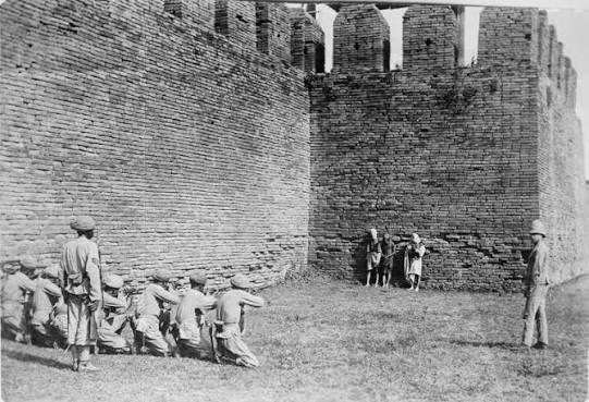 Execution of Burmese dacoits at Mandalay. A glass copy negative of a print showing three Burmese dacoits about to be executed by a British firing squad in front of the walls of
