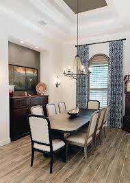 Dining room features a wood and metal oval dining table, a gray tufted settee under a window with a tan roman shade, a brown wooden klismos chair that boasts a white leather basketweave seat and a black shaded wall sconce. A La Carte Dining Room Window Treatments At Home Blinds Decor