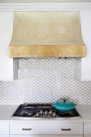 White kitchen island with black trim topped with a thick honed marble countertop illuminated by vintage glass pendants. A Gold Kitchen Vent Hood Is An Eye Catching Feature Over A Stainless Steel Stove Amongst White Quartz Countertops Kitchen Vent Hood Kitchen Vent Kitchen Hoods