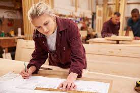 Carpenter With Apprentice Looking At Plans In Workshop How To Plan Apprentice Wood Crafting Tools