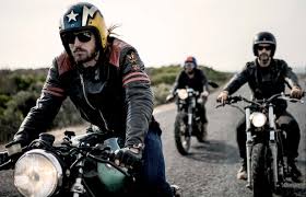 Three Men Wearing Open Face Crash Helmets And Sunglasses Riding Cafe Racer Motorcycles Along Rural Road