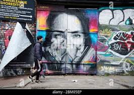 London May 2020 The Covid-19 pandemic. Brick Lane, Tower Hamlets. A man  walks his dog in front of Zabou street art of a woman wearing a gasmask  Stock Photo