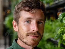 masculino com curto Castanho cabelo e luz barba vestindo Sombrio verde  camisa, em pé perto exuberante vegetação. ele parece diretamente às a  Câmera com uma relaxado expressão, dentro de casa, suavemente aceso