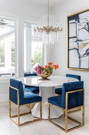 A Round White Dining Table Finished With Gold And Blue Sapphire Dining Chairs Under A Brass Dining Room Inspiration Modern Dining Room Dining Room Paint Colors