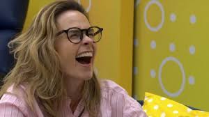 A mulher com óculos e cabelo loiro está rindo com a boca aberta, usando uma camisa listrada rosa e branca.