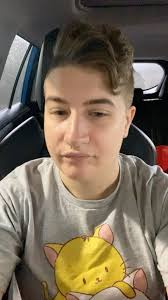 Video description: Ace is sitting in their car, which is parked in a garage  enclosure. Their hair swoop is gentle, yet stylish today, and they’re  wearing a grey T-shirt with a yellow cat head peeking ...