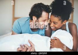 Happy, loving and funny interracial couple smiling and laughing while lying  in bed and sharing a tender moment. Carefree husband and wife relaxing and  having fun while bonding in the bedroom Stock