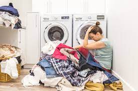 An Image of a woman sitting with a washing machine behind her and clothes on the floor , she look frustrated