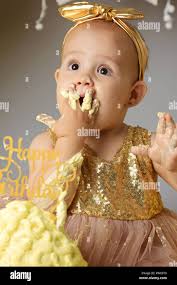 Small Sweet Baby Girl In A Golden Dress With A Bow On Her Head Trying A Jazzy Jelly Cake From A Cream Studio Shot Of A Birthday On A Gray Background Surrounded