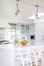 White Wishbone Stools Accented With Pink Cushions Sit At A Light Gray Island Topped With A White Quartz Countertop White Kitchen Island Kitchen Counter Stools Tan wishbone counter stools sit at a black kitchen island positioned a white quartz waterfall countertop illuminated by black industrial pendants hung from a wood beam over an industrial cooktop.