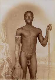 A young African man posing naked, holding a stick and a piece of cloth, in  front of a painted backdrop. | Wellcome Collection