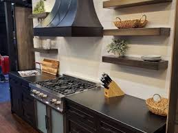 A steel hood accented with brass straps is mounted between stacked white shaker cabinets above a stainless steel oven range fixed beside a stainless steel microwave against a marble slab backsplash between white cabinets. Best Custom Range Hoods Available In Toronto Ontario Canada