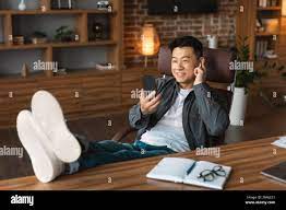 Glad confident adult korean male in wireless headphones has video call,  looks at webcam at workplace with laptop Stock Photo - Alamy