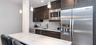 Sophisticated basement wet bar features gray and gold barstools that compliment a blue shaker wet bar island boasting white quartz countertop, facing a wall with white glass brick pattern tiles with mounted tv, flanked by dark blue shaker wet bar cabinets accented with brass hardware. 10 Top Trends In Basement Wet Bar Design For 2021 Sebring Design Build