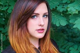 Stunning young woman with gray eyes and an ombre hair mix of dramatic  colors in tones of crimson red and orange, giving her a dazzling look  against green forest bush leaves. Stock