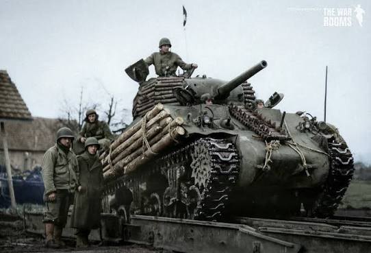 An M4A3 Sherman displays field modified protection with logs and spare track links as its crew prepares for operations in the European Theater during World War II.