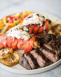 Well done style t bone steak and lobster plate on table beside roasted vegetables side dish and glass of water. Surf And Turf Steak And Lobster Tails Ahead Of Thyme