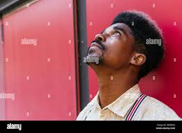 Tranquillo maschio afroamericano con capelli ricci appoggiati al rosso muro  di edificio e guardando in su mentre sogna Foto stock