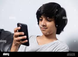 A cute young Indian boy sitting on his couch as he checks his mobile phone,  surfing the internet and social media. Video call with friend Stock Photo -  Alamy