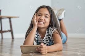 Smiling Little Girl Holding Mobile Phone Lying On Floor. Portrait Of Cute Indian  Girl With Toothy