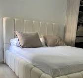 Esta cama presenta un diseño contemporáneo y elegante, con una estructura  tapizada en tela de textura suave, probablemente tipo bouclé o similar. ,  Sus formas acolchadas y voluminosas tanto en la base ...