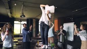 clow motion of joyful young lady dancing on table at office party with pile of paper and throwing documents while her team is laughing and dancing around her