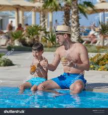 Dad And Son Are Sitting On The Side Of The Swimming Pool Drinking Lemonade And Making Like Gestures They Are At The Su Swimming Pools Swimming Summer Holiday