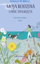 Okładka ksiażki Geralda Durrela pt. Moja rodzina i inne zwierzęta. Na okładce widzimy morze, plażę, kwiaty, kobietę idaca z psem na spacer.