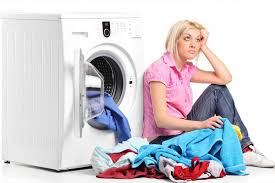 An Image of a woman sitting with a washing machine behind her and clothes on the floor , she look frustrated