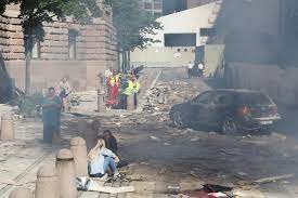 Omgitt av støv, røyk og bygningsrester står en ødelagt bil. 22 Juli Terrorisme 22 Juli Innspilling I Regjeringskvartalet