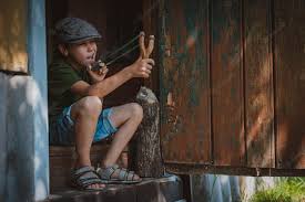 Niño niño en una gorra apunta con una honda para disparar a un objetivo. juega como un niño en el pueblo de vacaciones. | Foto Premium