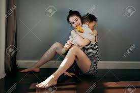 本当の瞬間、子供の息子は母の日に笑顔の母親のガーベラの花を与えます。幸せなお母さんは床に座っている男の子を抱きしめます。本物の家庭生活、ハードライト。の写真素材・画像素材  Image 203614154