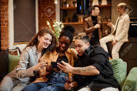 ホームパーティーでビデオチャットでコミュニケーションをとる3人の幸せな異文化の女の子が、リビングルームのソファに座って男たちと話しているの写真素材・画像素材  Image 191324467